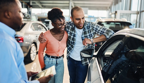Couple inspecting a car with a dealership representative in a showroom while discussing the vehicle's features.