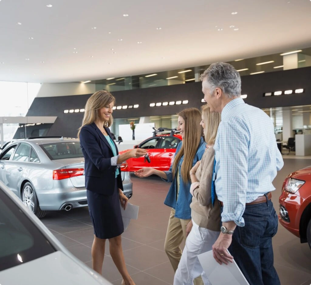 Sales representative handing car keys to a customer while two other family members stand beside them in a bright car dealership showroom with several vehicles on display.