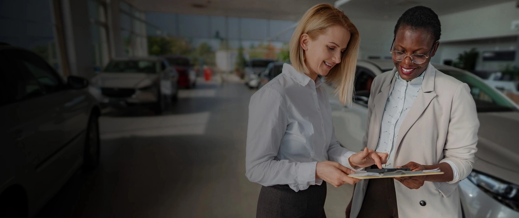 Two women shaking hands in a car dealership showroom, with new vehicles displayed in the background.