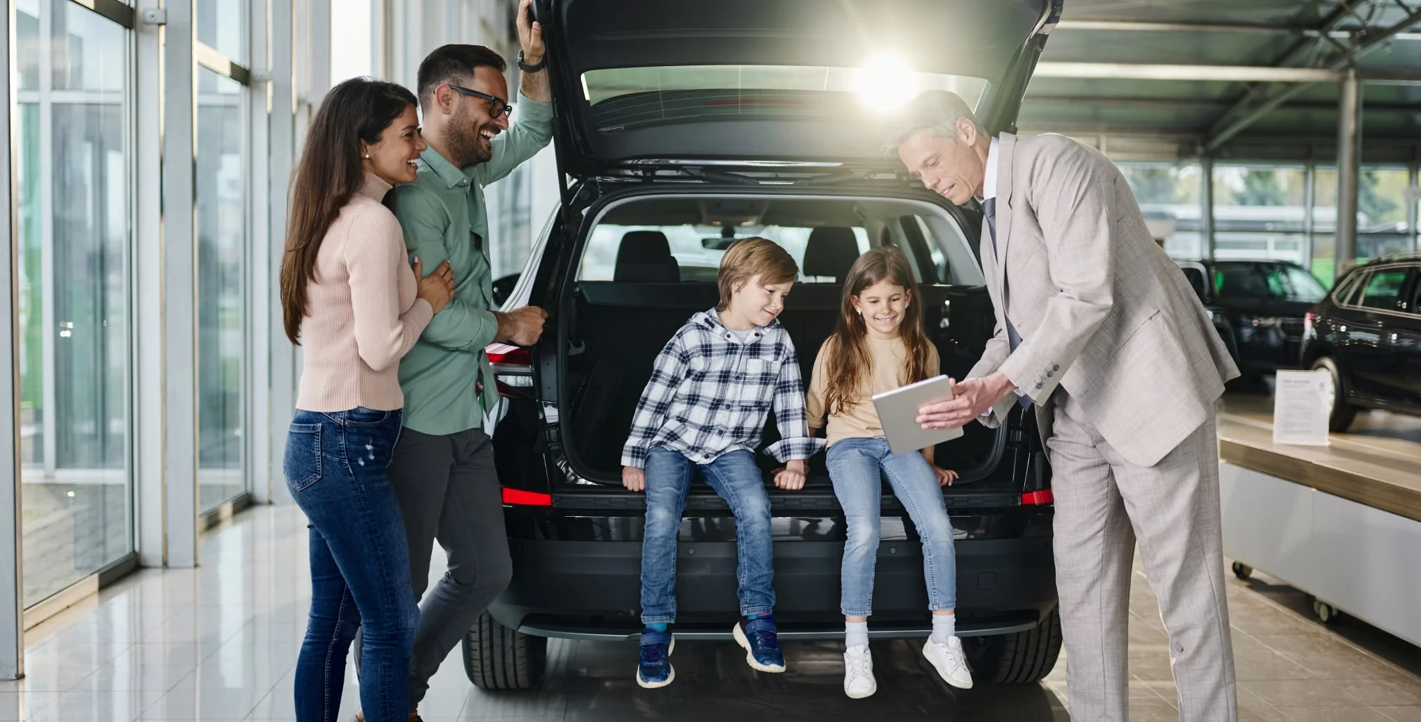 Two children sit in the open trunk of a car looking at a tablet with a dealership representative while their parents stand behind them